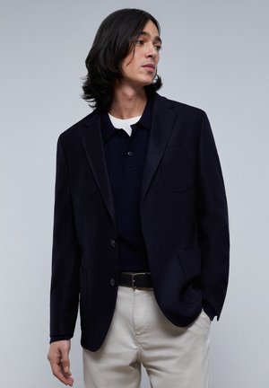 Blazer blu navy realizzato in tessuto testurizzato, dotato di una tasca sul petto, revers scollati e due bottoni, abbinato a una polo scura e a pantaloni chiari.
