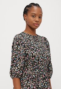 Svart blomstret kjole med korte pufferm, med små rosa, blå og kremfargede blomster på en mørk bakgrunn; stoffet ser lett ut.