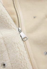 Crème-kleurige jas met een gestructureerde, fleece-achtige interieur en een gladde buitenkant. Inclusief een zilveren rits en knoopdetails.
