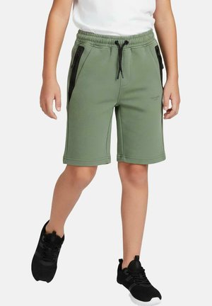 Kind draagt groene short met trekkoord en zwarte ritszakken, een wit t-shirt en zwarte sneakers, staat met één been licht gebogen.