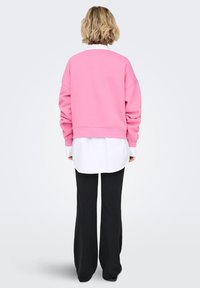 Ροζ κοντή φούτερ με μακριά μανίκια, φορεμένη πάνω από ένα άσπρο πουκάμισο με κουμπιά, σε συνδυασμό με μαύρο παντελόνι φαρδιάς γραμμής, παρουσιάζοντας ένα στιλ με στρώσεις.