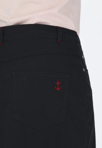 Pantaloni corti neri con tasca posteriore decorata da ricamo di un'ancora rossa, cuciture a contrasto e una texture liscia. Passanti per cintura visibili e dettaglio del bottone.
