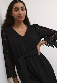 Abito nero con scollo a V, tessuto fluente e maniche a tre quarti con dettagli in pizzo nero intricati. Include una cintura coordinata.
