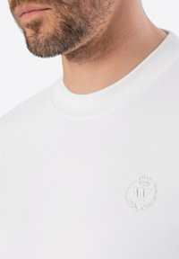 Bílý tričko s krátkým rukávem s kulatým výstřihem a vyšívaným logem s vavřínovými akcenty na hrudi. Hladká textura.