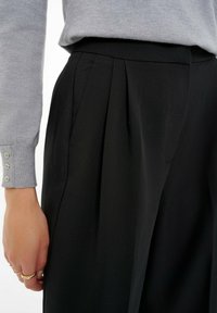 Pantalones negros con pliegues, de textura suave, corte entallado y cinturilla plana, combinados con un suéter gris con detalles de botones.