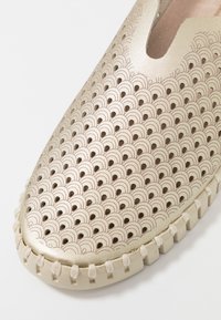 Zapato tipo slip-on en dorado metálico con un patrón texturizado y ondulado. Presenta una suela acolchada y un detalle trenzado alrededor del borde.