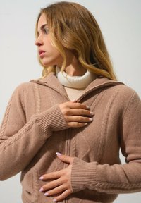 Beige Strickcardigan mit Kapuze, mit einem Zopfmuster und gerippten Bündchen. Getragen über einem weißen Rollkragenpullover.