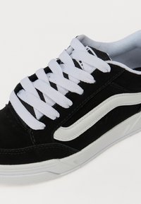Fekete velúr sneaker fehér gumitalppal és fehér fűzővel. Fehér oldalsávval és textúrázott panelek részletekkel rendelkezik.