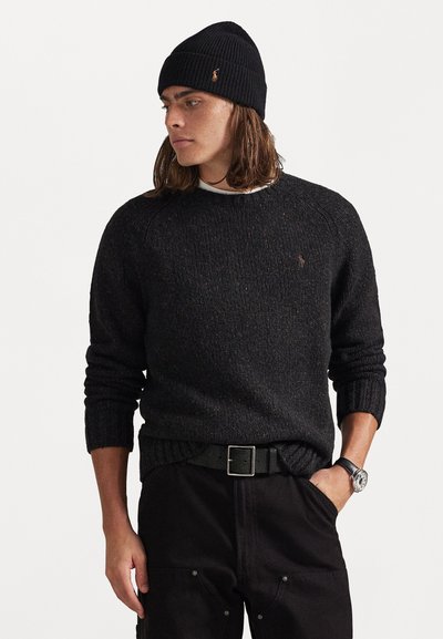Black wool sweater with ribbed cuffs and crew neckline, paired with a black beanie. Slim black pants and silver wristwatch complete the look.