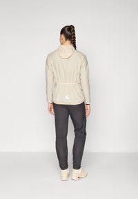Svijetlosmeđi zip-up jakna s kapuljačom, elastičnom strukom i stražnjim trokutastim logom, u kombinaciji s tamnim hlačama i bež tenisicama.