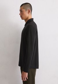 Zwarte lange mouw poloshirt met een kraag, slim fit en een gladde, zachte textuur. Draagt bij olijfgroene broeken, tegen een neutrale achtergrond.