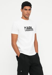Valge puuvillane T-särk, mille esiküljel on must "KARL LAGERFELD" logo. Lühikesed varrukad ja ümar kaelus, kombinatsioonis mustade cargo-pükstega.