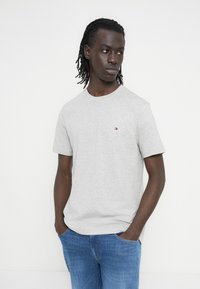 Γκρι βαμβακερό μπλουζάκι με κοντά μανίκια, στρογγυλή λαιμόκοψη και μικρό κεντητό λογότυπο στο στήθος. Συνδυάζεται με μπλε τζιν παντελόνι.