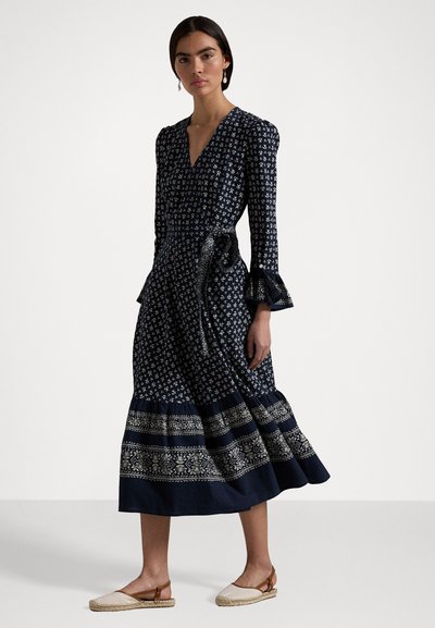 Nainen, jolla on päällään tummansininen midi-mekko, jossa on kukkakuviota, röyhkeä hihat ja vaaleanbeige espadrillo-kengät, seisoo yksinkertaista taustaa vasten.