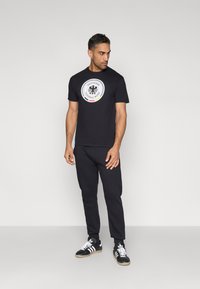 Fekete póló, amelyen kerek, fehér logó látható, a Német Labdarúgó Szövetség emblémájával; hozzáillő fekete melegítőnadrággal és fekete sportcipővel viselve.