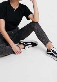 Camiseta negra de manga corta combinada con jeans ajustados de cintura alta en gris oscuro y zapatillas negras con detalles blancos. Pose sentado sobre un fondo claro.