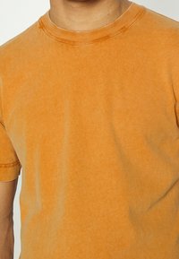 Krátký rukáv oranžového trička vyrobeného z měkké bavlny, s klasickým kulatým výstřihem a jemnou strukturou. Obsahuje zesílené švy kolem límce.