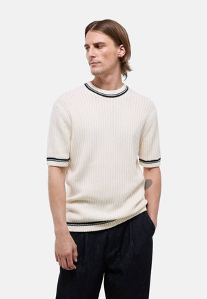 Homme aux cheveux mi-longs portant un pull en maille blanc à manches courtes avec des rayures noires, les mains dans les poches d’un pantalon foncé, regardant vers la gauche.