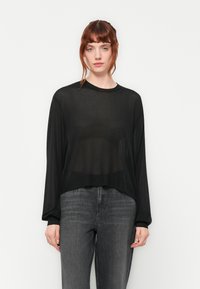 Haut long à manches transparentes noir avec un col rond, coupe décontractée et texture subtile, associé à un jean gris foncé.