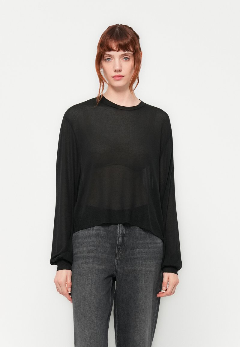 Haut long à manches transparentes noir avec un col rond, coupe décontractée et texture subtile, associé à un jean gris foncé.