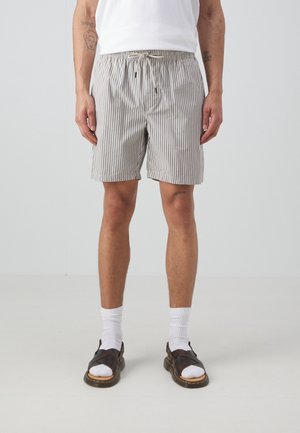 Homme portant un short beige et blanc à rayures verticales, des chaussettes blanches, des sandales marron et un t-shirt blanc, debout devant un fond uni.