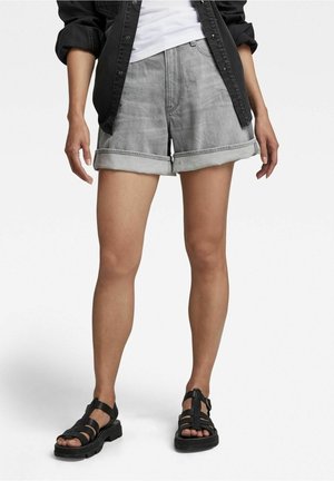 Persoon draagt grijze opgerolde spijkershorts, zwarte sandalen met open tenen, wit overhemd en zwart knoophemd met opgerolde mouwen.