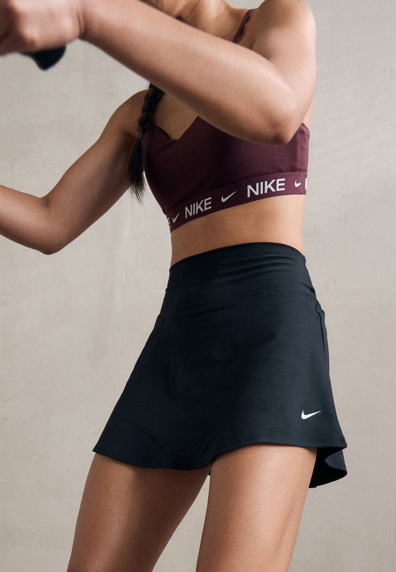Femme portant un soutien-gorge de sport Nike bordeaux et une jupe de sport Nike noire, photographiée de la moitié du torse à la moitié de la cuisse, sur un fond neutre.