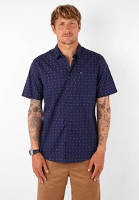 Chemise à manches courtes bleue marine avec un motif géométrique, dotée d'une poche poitrine et d'une texture de tissu lisse. Mains tatouées visibles.