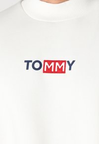 Bijela dukserica s rebrastim okruglim izrezom, s vezanim "TOMMY" logotipom u tamnoplavoj i crvenoj boji. Mekana tekstura, ležeran kroj.
