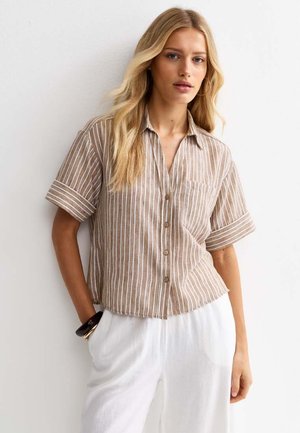 Blondynka w beżowo-białej koszuli w paski z krótkim rękawem zapinanej na guziki oraz białych spodniach, stojąca z jedną ręką w kieszeni.