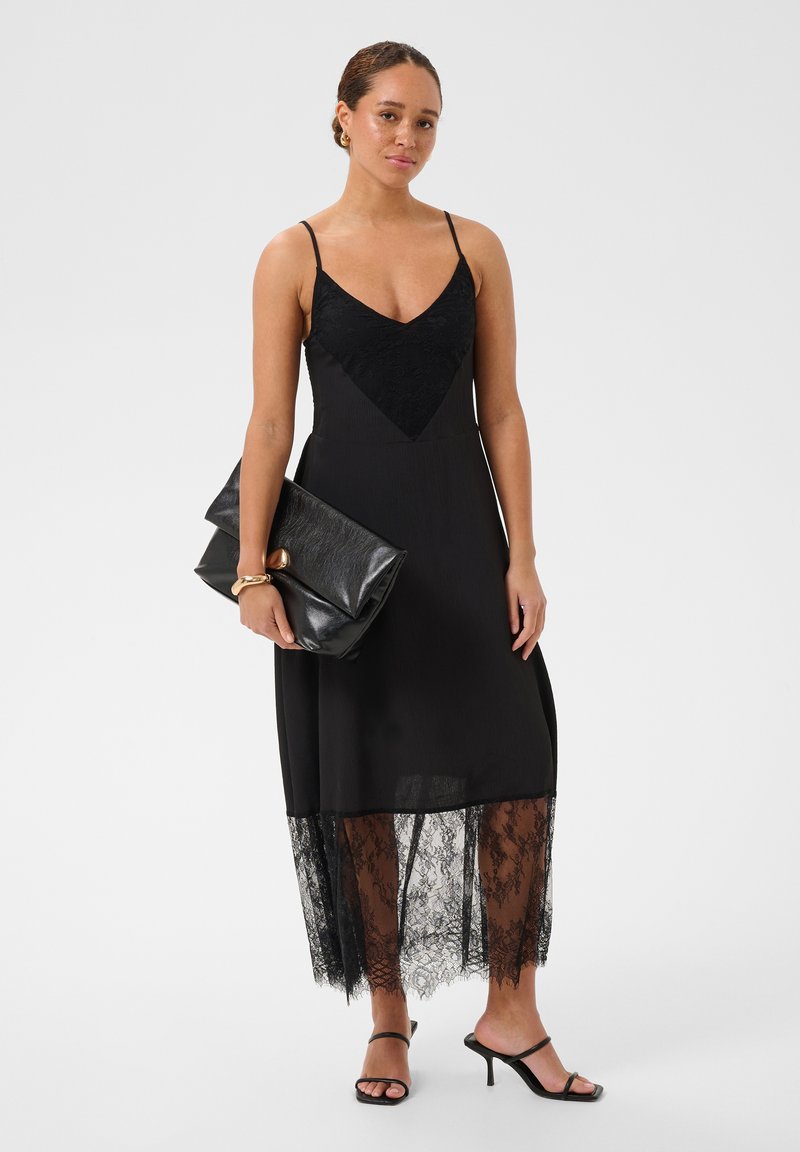 Femme en robe midi noire bordée de dentelle, tenant une grande pochette noire, portant des sandales à talons noires et un bracelet en or.
