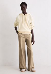 Lichtgele hoodie, ruime pasvorm; gecombineerd met op maat gemaakte, beige wijde broek en lichtgekleurde instapschoenen.