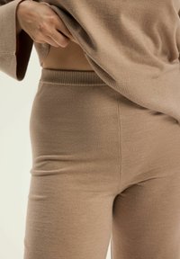 Beżowy sweter dziane układa się na dopasowanych spodniach, które mają prążkowany pas i gładką fakturę, podkreślając bezszwowy design.