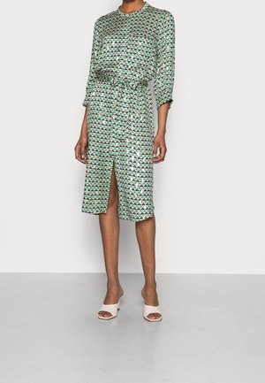 Vrouw draagt een knie-hoog zijden jurk met een groen en marineblauw driehoekspatroon, een frontale split en beige open-toe sandalen met hak.