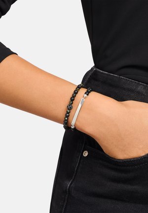 Arm mit zwei Perlenarmbändern, eines mit einer Silberstange, ruhend auf einer schwarzen Jeans-Tasche vor einem schwarzen Ärmelhintergrund.