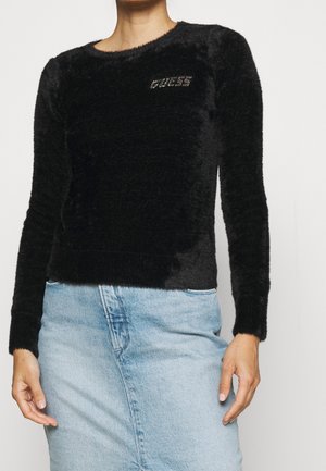 Femme portant un haut noir à manches longues duveteux avec "GUESS" en strass sur la poitrine et une jupe en jean bleu clair. Haut du corps visible.