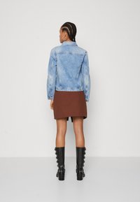 Veste en denim bleu clair avec une coupe ajustée, un empiècement au dos et des poignets boutonnés, associée à une jupe marron longueur genou et des bottes noires à hauteur de genou.