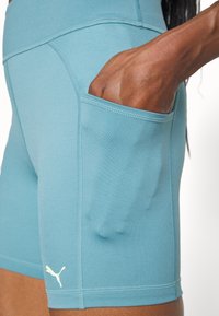Main dans la poche d’un short de sport Puma bleu clair avec coutures visibles et logo sur la cuisse.