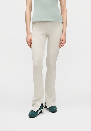 Žena v béžových rozšírených nohaviciach s prednými rozparkami a tmavozelených teniskách značky Puma, stojaca pred jednoduchým bielym pozadím.