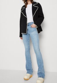 Musta kaksirivinen takki, jossa on valkoista kontrastipistoa, yhdistettynä vaaleansinihin leveälahkeisiin farkkuihin ja kermaisiin puukenkiin.