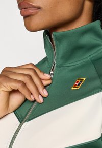 Grønn zip-jakke laget av glatt stoff, med høy krage, hvit aksentpanel og en fargerik brodert logo på brystet.
