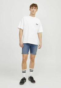 Camiseta blanca de gran tamaño con texto impreso, pantalones cortos de mezclilla azul con puños doblados, zapatillas negras y calcetas blancas hasta la rodilla con rayas negras.