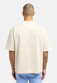 Beige oversized t-shirt featuring embossed text and a winged design on the back, with short sleeves and a slightly textured finish.