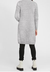 Gray knitted long cardigan with ribbed hem, worn over black slim-fit pants and white ankle boots. Texture appears soft and cozy.