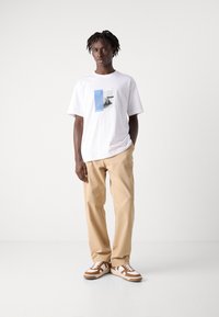 Hvit t-skjorte med korte ermer som har et grafisk design med blå detaljer, kombinert med beige bukser og brune og hvite sneakers.