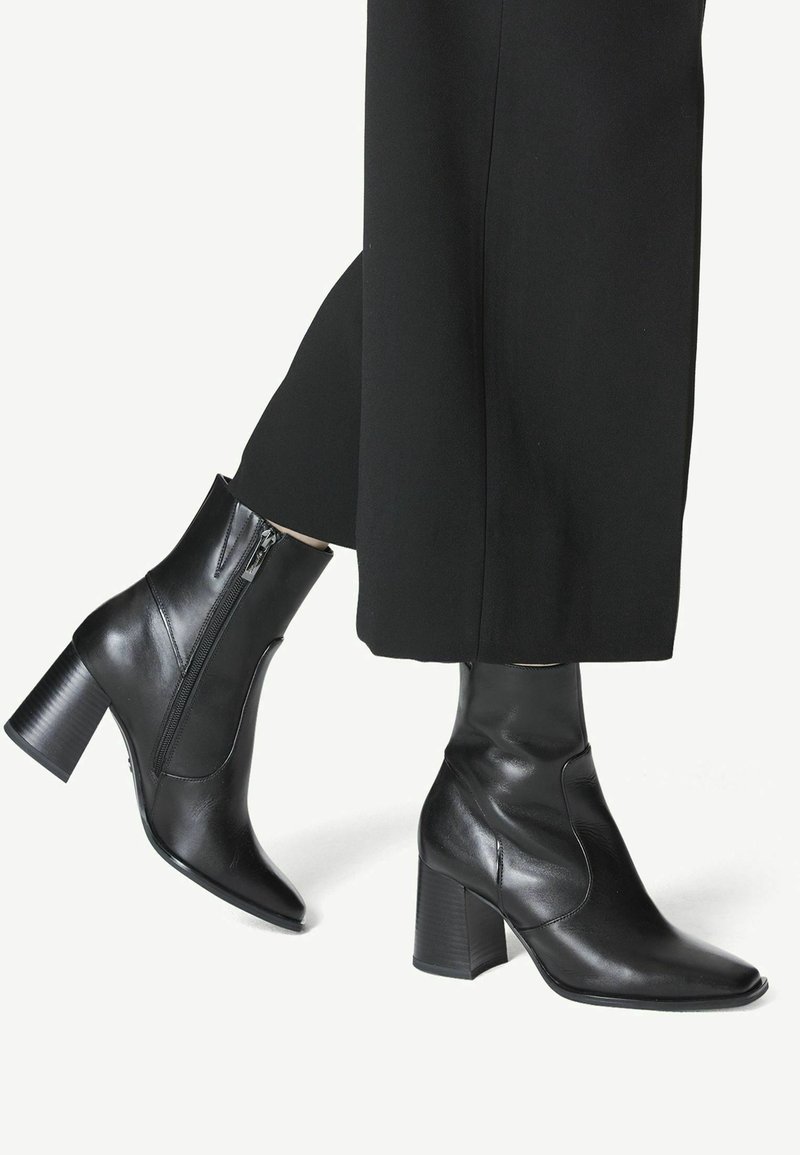Bottes montantes en cuir noir avec talons carrés et fermetures éclair latérales, portées avec un pantalon noir raccourci sur un fond clair.