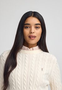 Crème kabelgebreide trui met een ronde halslijn, gedragen over een lichtroze gingham shirt, met een klein geborduurd logo op de borst.
