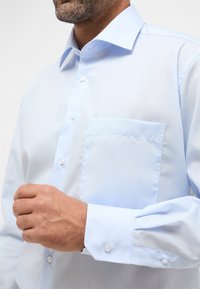 Camicia azzurro chiaro con colletto a punta, taschino sul petto e maniche con polsini. Tessuto liscio con una texture uniforme.