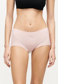 Γυναίκα φοράει ανοιχτό ροζ βαμβακερά boyshorts με τη λέξη «Friday» τυπωμένη μπροστά και μαύρο κοντό τοπ, στέκεται μπροστά σε λευκό φόντο.