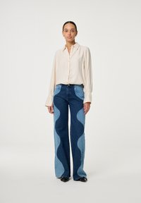 Beige overhemd met lange mouwen en gestructureerde manchetten, gecombineerd met blauwe wijde jeans met golvende paneelontwerpen in twee tinten denim.